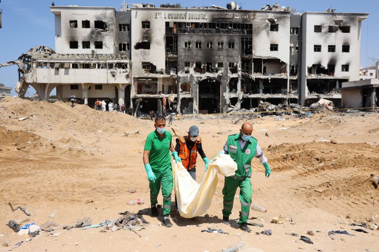 people wearing green and orange uniforms carry a body in a white bag outside a destroyed building