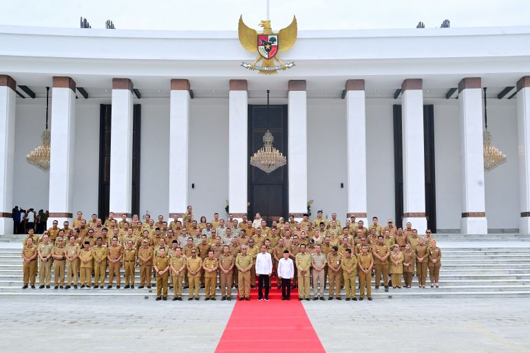 Indonesia's cabinet standing outside the new presidential palace in Nusantara. The buiding is behind them. It looks vast with tall white columns. The Indonesian insignia is on the roof