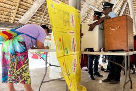 A woman voting in the Kirbati election. She is bent over a table making her choice. Two policemen are moving one of two ballot boxes nearby