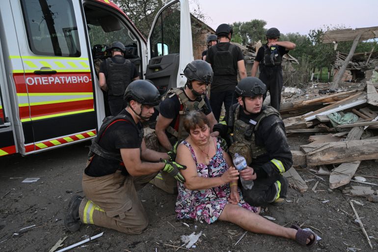 A Ukrainian woman being helped by resue teams after she fainted. An ambulance is behind her. There is lots of rubble around.