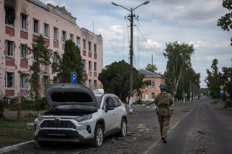 Soldier walks past a damaged building and a silver car