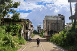 A street in Marawi. It is lined by ruined and abandoned buildings. Plants are growing out of them. Two people are walking down the middle of the street away from the camera. There are no cars and noone else is around.